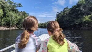 duas crianças, de costas para câmera, em barco em rio da Amazônia, nas margens se veem muitas árvores. É um dia de sol com poucas nuvens.