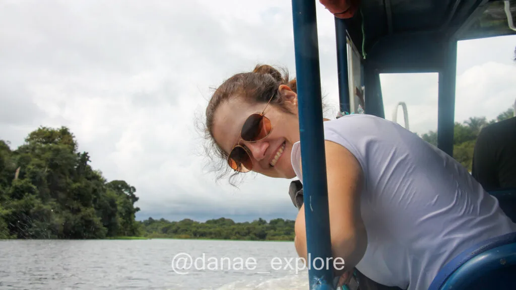 I, a white woman, wearing sunglasses and a white t-shirt, leaning over the side of a boat, looking back at the camera, as the boat sails through the waters of the Rio Negro, near Anavilhanas National Park