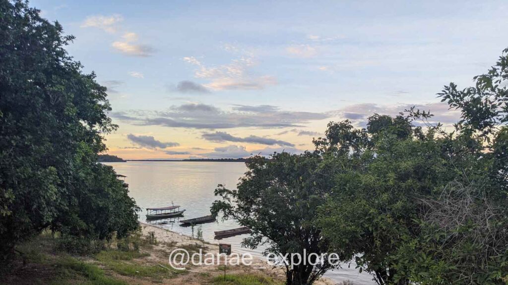 Por-do-sol no Rio Negro, se vê o rio, algumas ilhas com vegetação no horizonte, em primeiro plano há ávores, uma pequena praia com alguns barcos