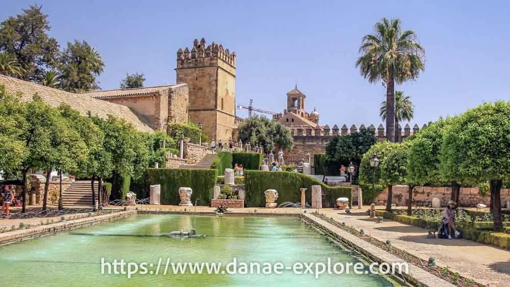 Alcazar de los Reyes Cristianos, Cordoba, Espanho. Em primeiro plano um espelho d'água e jardim, ao fundo uma torre, em dia de sol e céu azul