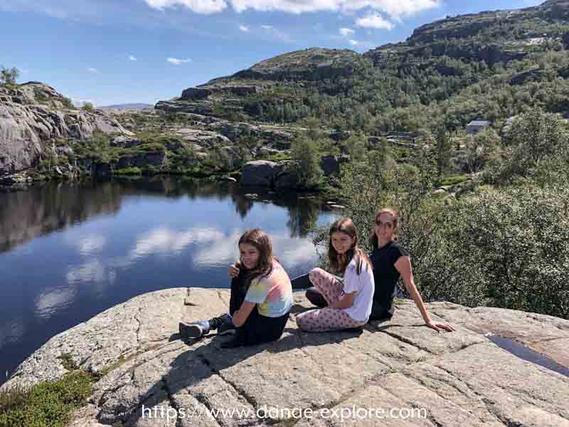 trilha para Preikestolen, Pedra do Púlpito, Pulpit Rock, Noruega. Mãe e duas crianças sentadas na pedra, olhando para a camera, em dia de sol com céu azul e poucas nuvens