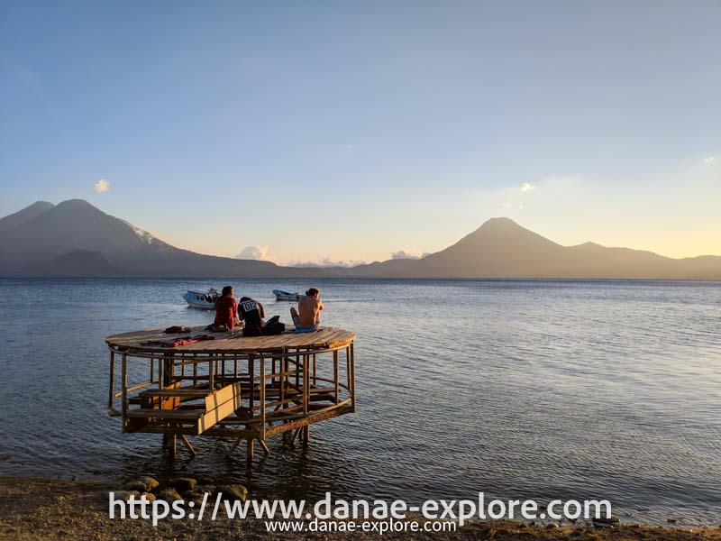 pessoas em deck de madeira observando o por-do-sol no Lago Atitlán, Guatemala. Há vulcões ao fundo e pequenos barcos no lado