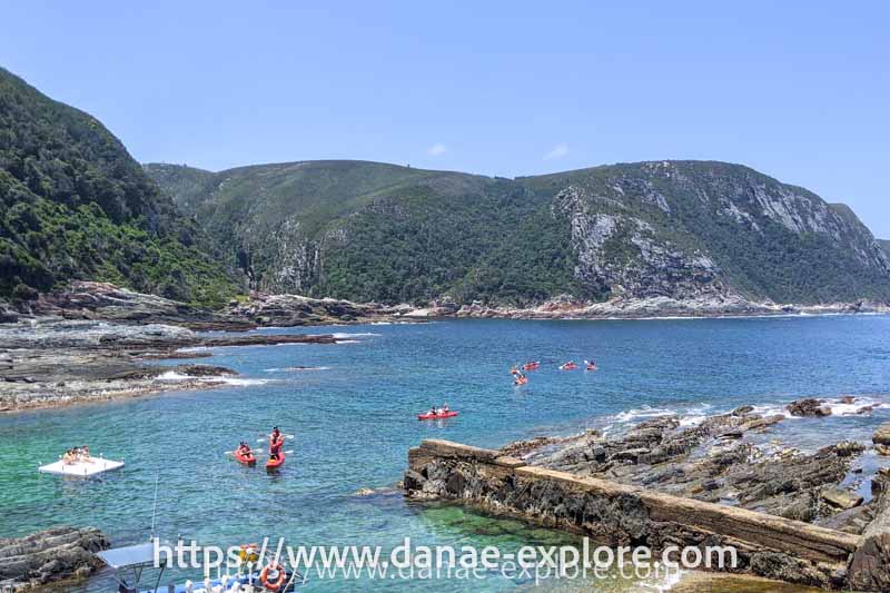 Pessoas em caiaque em praia com rochedos e montes com vegetação ao fundo, Tsitsikama Parque, África do Sul