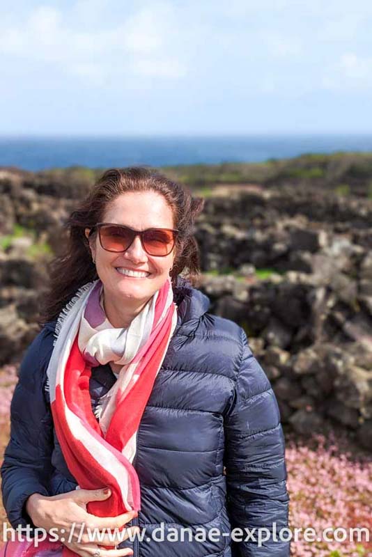 Moça branca de óculos escuros, blusa preta e  lenço em tons vermelho, sorrindo para câmera, ao fundo videiras em vinícola na Ilha do Pico, Açores, Portugal