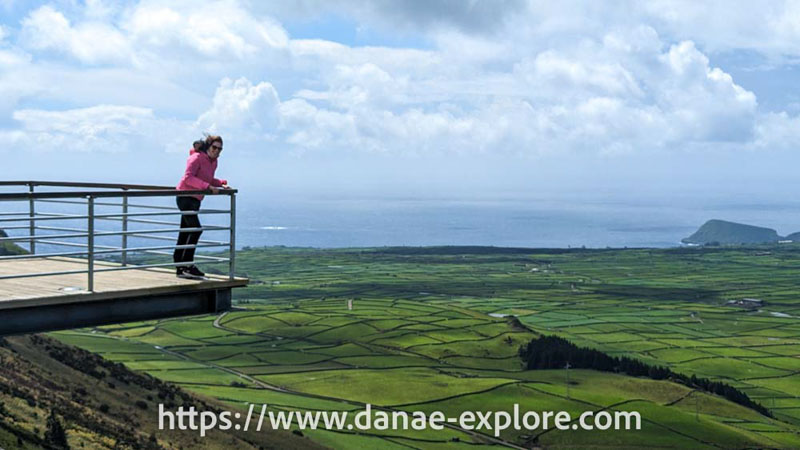 moça em calça preta e blusa rosa no Miradouro da Serra do Cume, Ilha Terceira, em dia nublado com vento. Na paisagem se veem campos verdes e o mar ao fundo