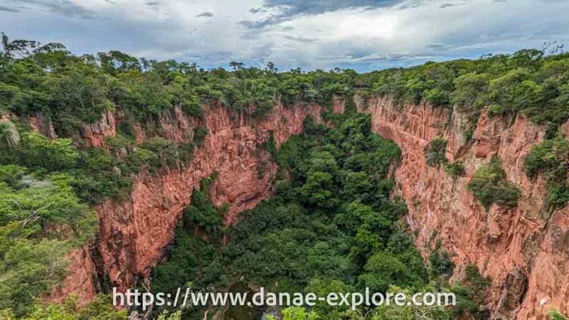 Vista ampla do Buraco das Araras, em Bonito - M.S. - é possível ver as paredes rochosas do buraco no solo, a vegetação verde e um pequeno lago ao fundo, sob um céu nublado.