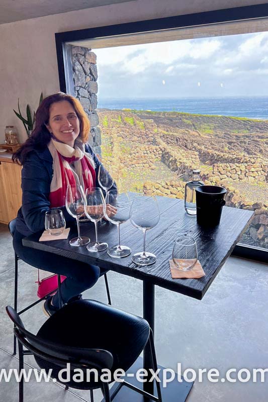 Fazendo degustação de vinhos na Ilha do Pico; moça em blusa preta e lenço em tons de vermelho sorri e olha para câmera, pela janela se veem os currais das vinhas do Pico. www.danae-explore.com