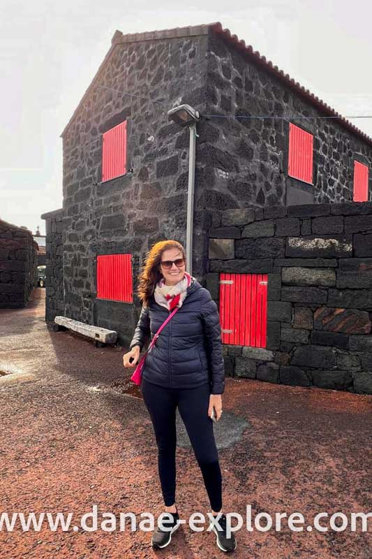 Moça de cabelos longos, blusa e calça preta em frente a casa em pedra preta vulcânica, argamassa branca, portas e janelas vermelhas, no vilarejo de Lajido, Ilha do Pico, Açores, Portugal - www.danae-explore.com