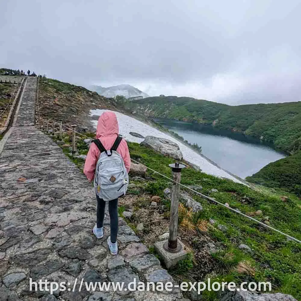 Murodo - Tateyama Kurobe Alpine Route, Girl in pink jacket and backpack walking, on a cloudy day, next to a lake with remnants of snow on the slopes