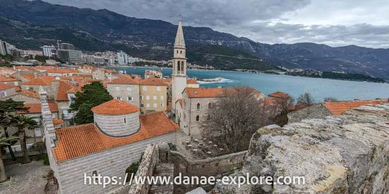 Vista do alto da cidade antiga de Budva, Montenegro. Prédios baixo em pedra clara, há uma torre de igreja e à direita se avista o mar. 
