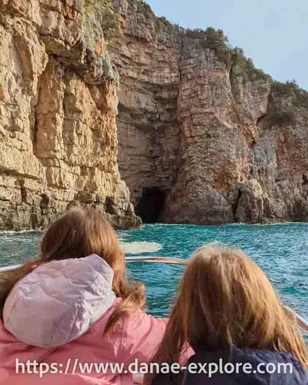 Duas garotas, de costas, em um pequeno barco, mar azul, o barco ruma a uma pequena entrada na parede rochosa, formando uma pequena caverna - Blue Cave, Montenegro