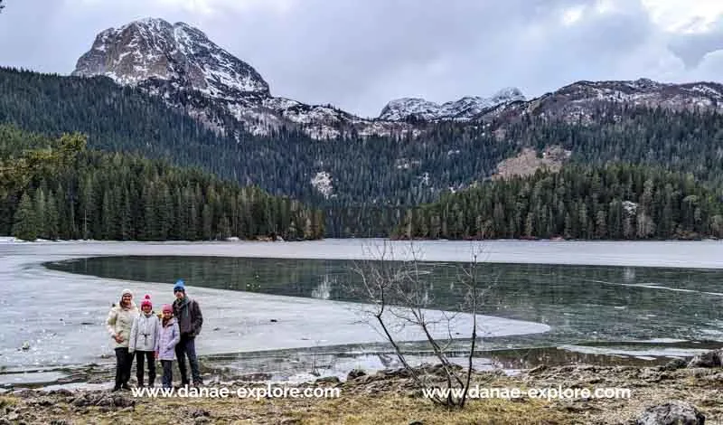Família (2 adultos e 2 crianças) em frente a um lago, com montanhas nevadas e florestas de pinheiros ao fundo. É inverno e todos vestem ccasacos e gorros. Black Lake, Parque Nacional Durmitor, Montenegro. 