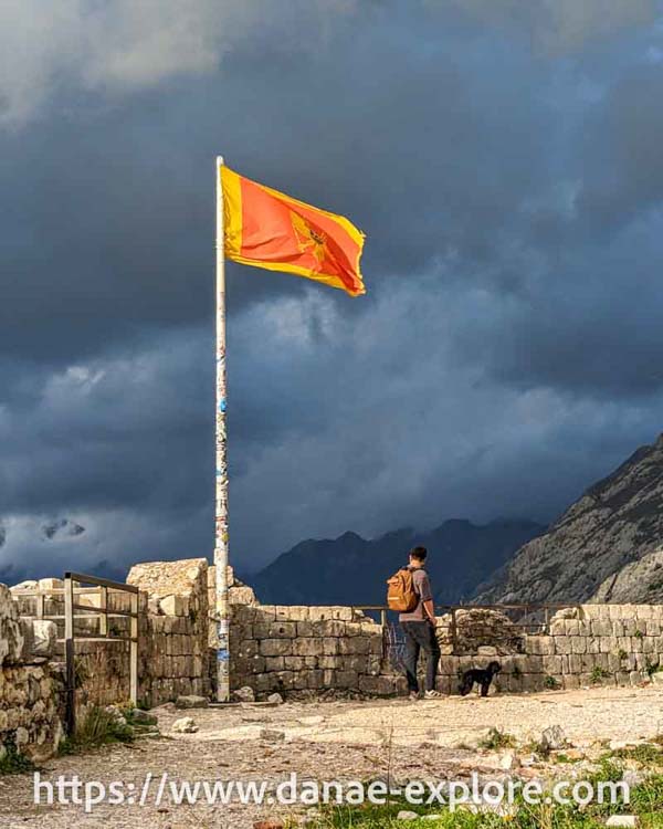 Bandeira de Montenegro em mastro no alto da Fortaleza de Kotor. Há um homem e um cachorro pequeno na foto e nuvens negras ao fundo.