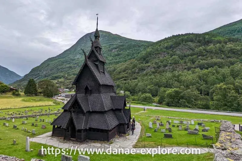 Igreja em madeira escura - Borgund Stave Church, ao fundo montanhas com vegetação verde em dia encoberto - parte de nosso post sobre quanto custa viajar na Noruega