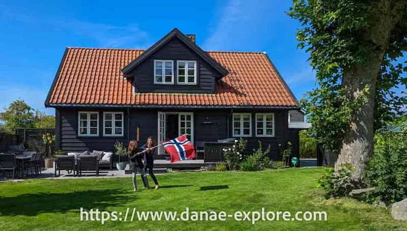 Duas meninas brincam com uma bandeira da Noruega no gramado verde do quintal de uma casa em madeira escura, com janelas brancas. É um dia com sol e ceu azul - quanto custa viajar na Noruega
