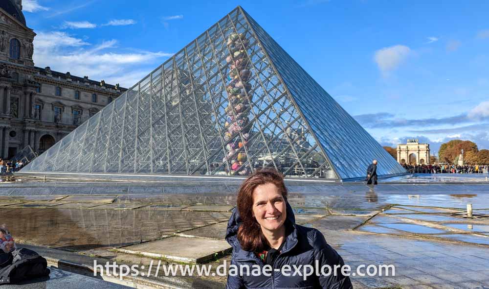 Moça branca de cabelos longos em blusa escura, em frente à pirâmide de vidro do Museu do Louvre, em dia de sol. Dicas de como planejar uma viagem de férias, www.danae-explore.com