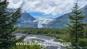 Paisagem natural do Alasca com árvores, rio pedregoso e Exit glaciar se estendendo pelas encostas das montanhas.
