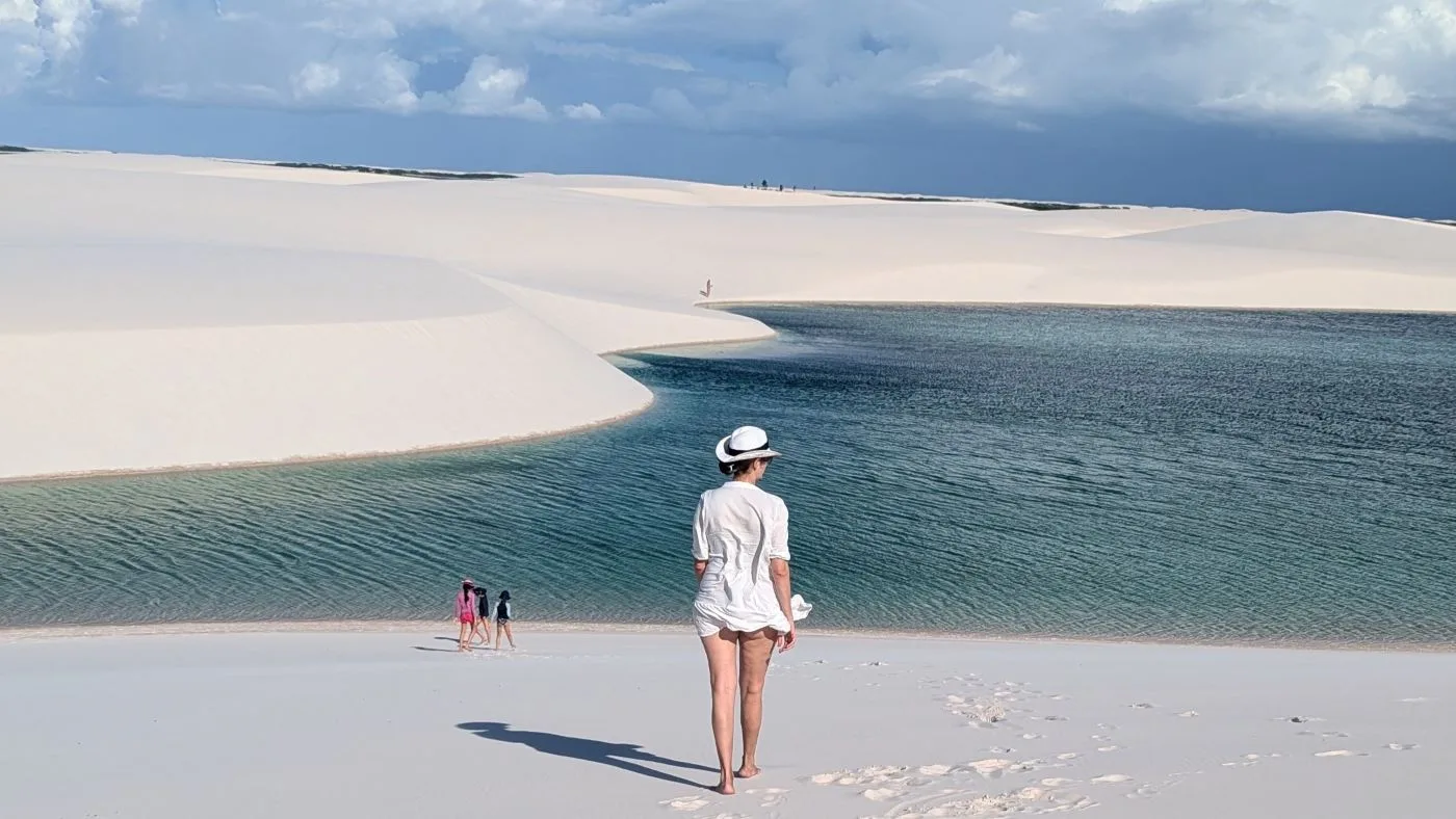 Mulher de chapéu branco caminha em direção a uma lagoa cristalina entre dunas nos Lençóis Maranhenses.