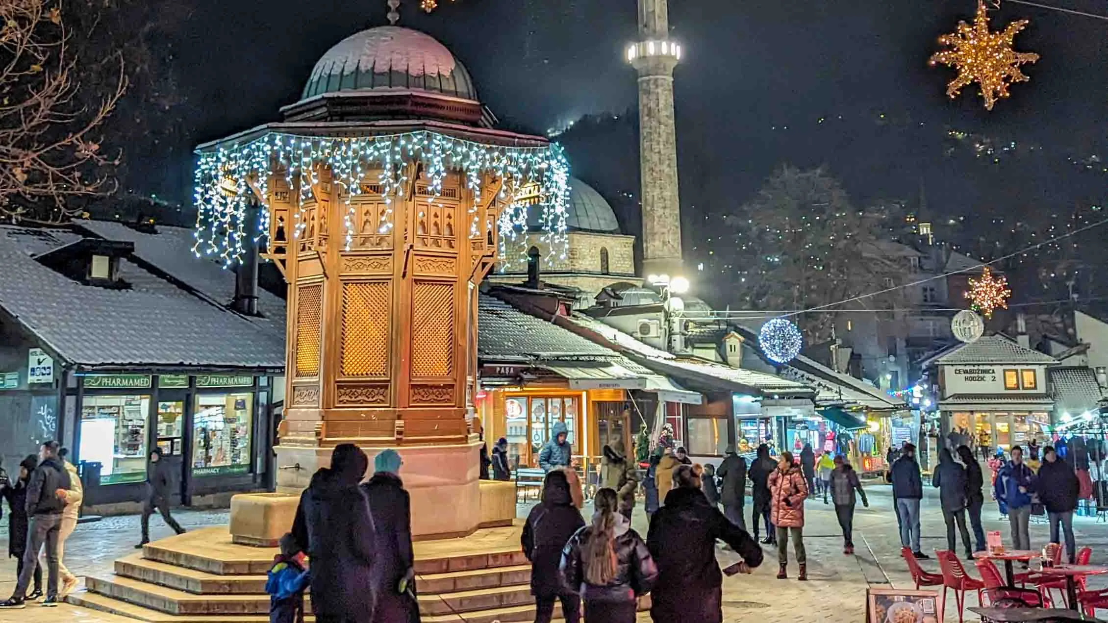 night view of Bascarsija in Sarajevo, with the Sebilj Fountain in the foreground; it is a winter night and there are many people walking along the streets