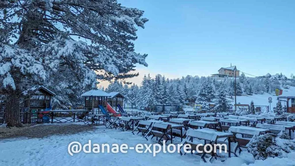 Outdoor tables and playground in Trebevic, covered in a thick layer of snow. In the background you can see pine trees, also covered in snow. It's a blue sky day.