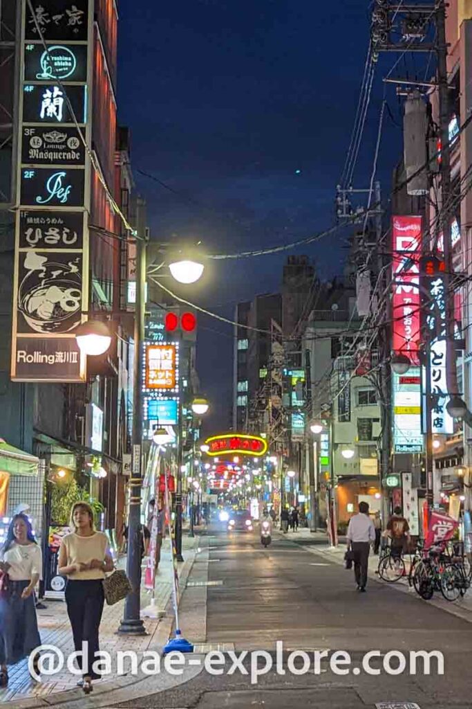 Hiroshima a noite, região de Hondori; Há diversas luzes neon e algumas pessoas andando pela calçada