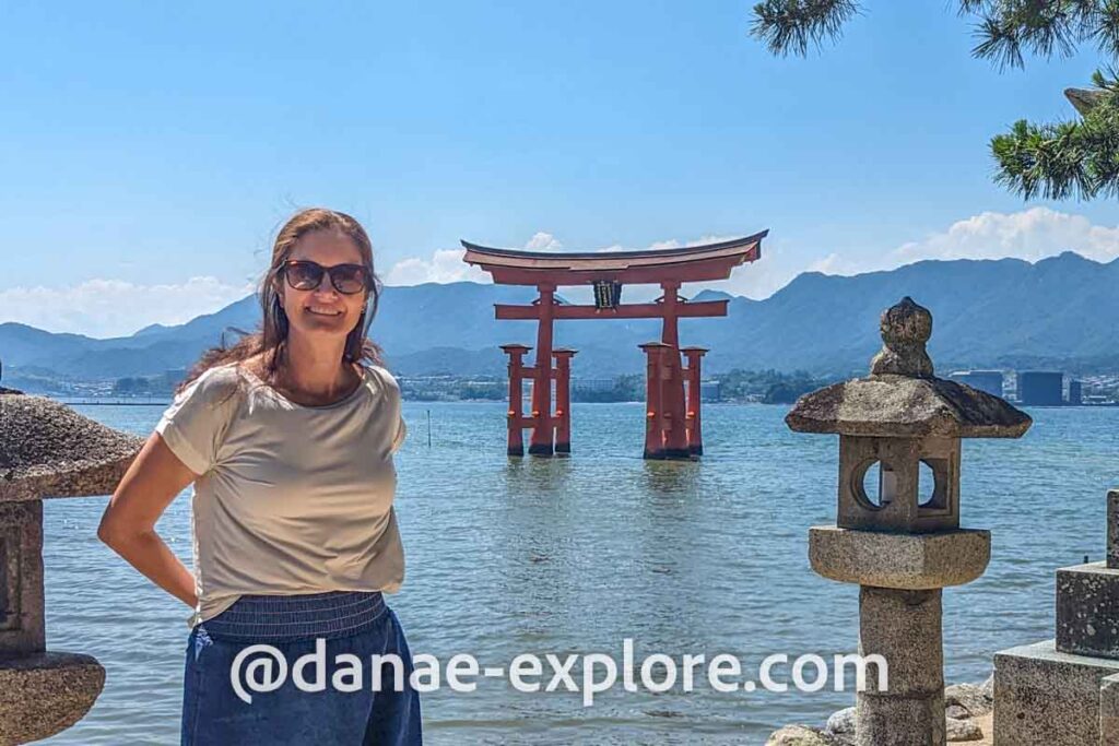 moça em blusa branca, saia jeans e óculos de sol, olhando para câmera em dia de sol e céu azul. Ao fundo a Torii flutuante do Santuário Itsukushima, em Miyajima