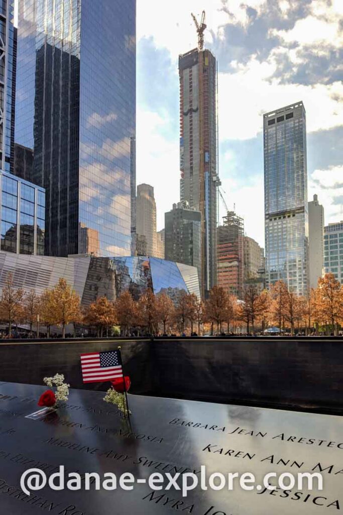 9/11 Memorial, em Nova Iorque, em dia de sol com algumas nuvens. Há uma homenagem a uma das vítimas, com flores e uma bandeira dos EUA ao lado de seu nome