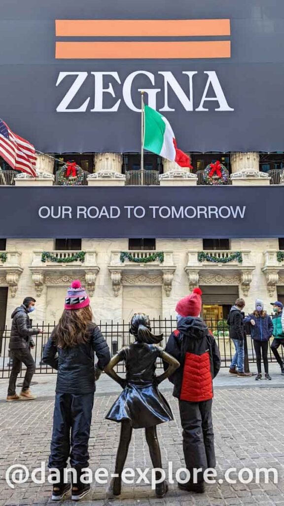 duas garotas, uma de cada lado da estátua da garota em frente a Bolsa de Valores de Nova Iorque. No prédio, há um grande painel onde se lê "Zegna - Our road to tomorrow"