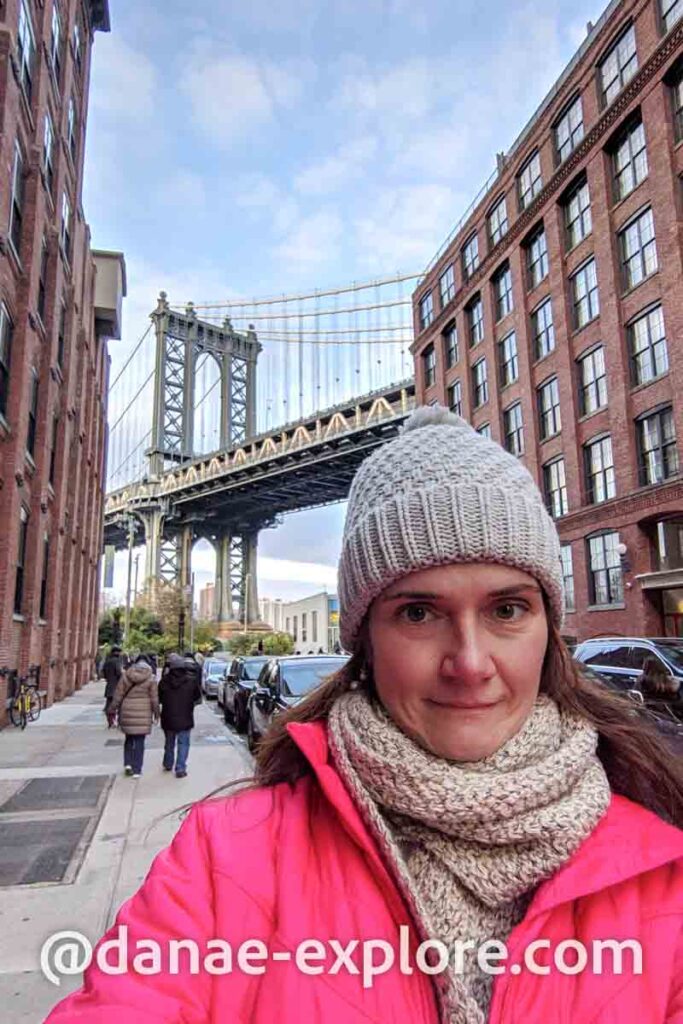 selfie de moça, em blusa vermelha, cachecol e gorro, no DUMBO em Nova Iorque. Ao fundo, se vê a Manhattan Bridge