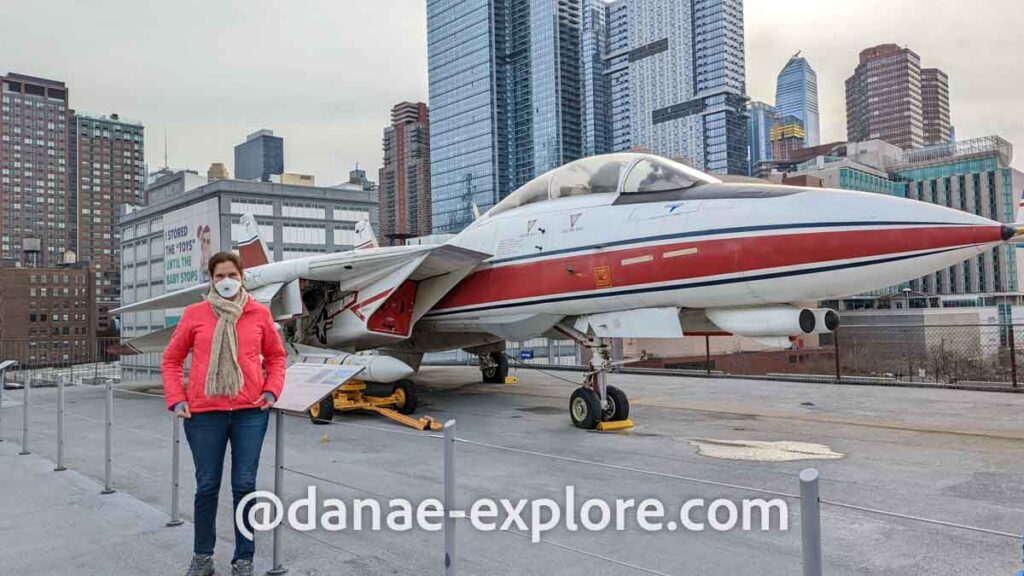 moça em blusa vermelha e calça jeans, com cachecol e máscara, em frente ao F-14 Tomcat no Intrepid Museum em NYC