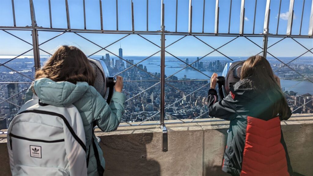 duas meninas em roupas de inverno, de costas para câmera, olham o skyline de Nova Iorque em binóculos no alto do observatório do Empire State Building