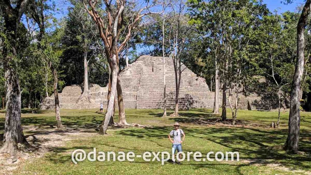 Homem posa para câmera em meio a algumas árvores, ao fundo se vê parte das ruinas maias de Cahal Pech, em Belize