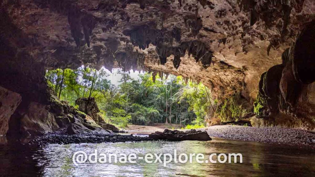 saída de caverna em Belize; há um rio dentro da caverna, estalactites no teto da cavena, e vegetação ao fundo, do lado externo da caverna