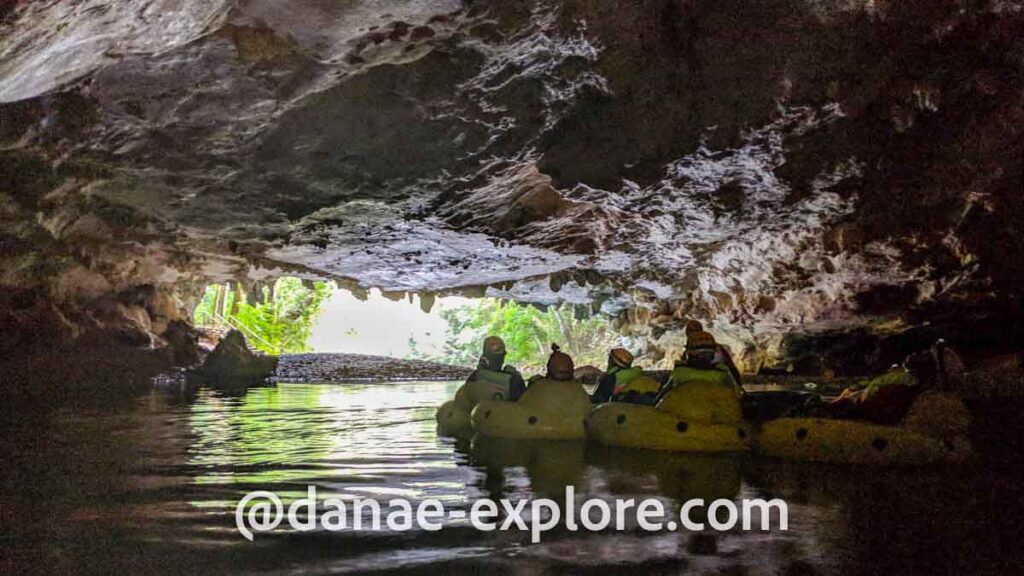 Cave tubing em Belize. Foto tirada de dentro de uma caverna, ao fundo se vê a saída da caverna, com vegetação. Em primeiro plano há um grupo de pessoas em bóias amarelas, e estalactites no teto da caverna