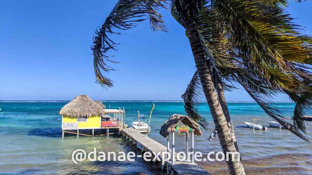 Caye Ambergris, Belize. Há um pier com uma casa de madeira amarela e teto de palha. O mar é azul turquesa. Se vê parte de uma palmeira em primeiro plano