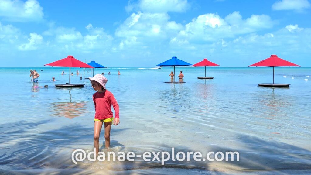 criança com camiseta vermelha e chapéu cor-de-rosa na praia. A água é azul clara e calma; ao fundo se veem guarda-sóis vermelhos e azuis na água, com algumas pessoas