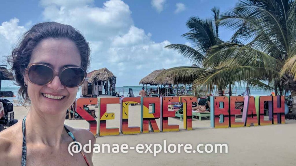 Selfie de moça branca com óculos escuros na praia, em frente a letreiro escrito "Secret Beach", uma das praias que visitamos em nosso roteiro de uma semana em Belize