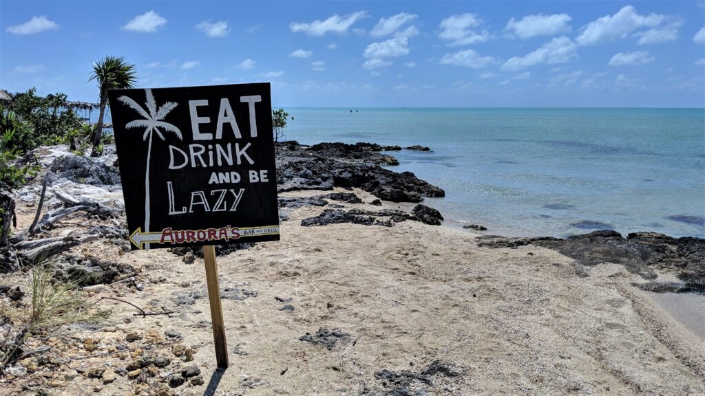 placa em praia de Belize onde se lê "eat, drink and be lazy". Ao fundo, o mar é azulo claro, sem ondas, e há poucas nuvens no céu