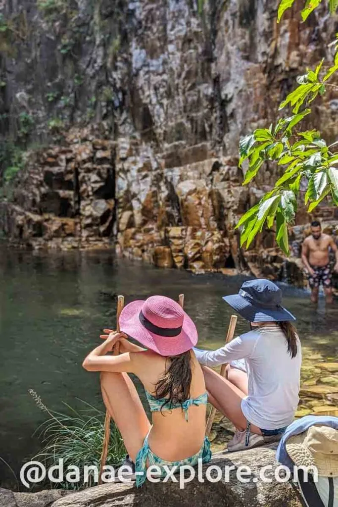 Duas meninas, de costas para camera, a sentadas a beira do poço de uma cachoeira da Cachoeira dos Dragões, em Pirenopolis.