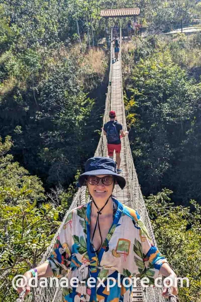Moça em chapéu azul e óculos de sol, sorrindo para câmera, em ponte pênsil na trilha da Cachoeira do Abade, em Pirenopolis, Goiás.