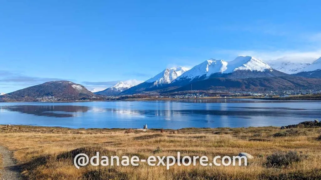 View of Ushuaia in autumn; there is a yellowish field in the foreground, a bay in the center and snow-capped mountains in the background