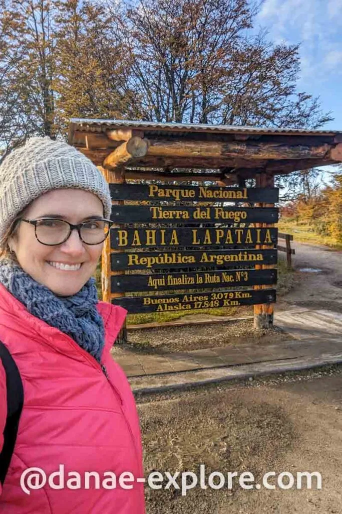 Selfie of a white girl with glasses, wearing a red coat, blue scarf and hat in front of a sign that reads: Tierra del Fuego National Park, Lapataia Bay, Argentine Republic, Here ends National Route No. 3 Buenos Aires 3.079 km, Alaska 17.848 km"