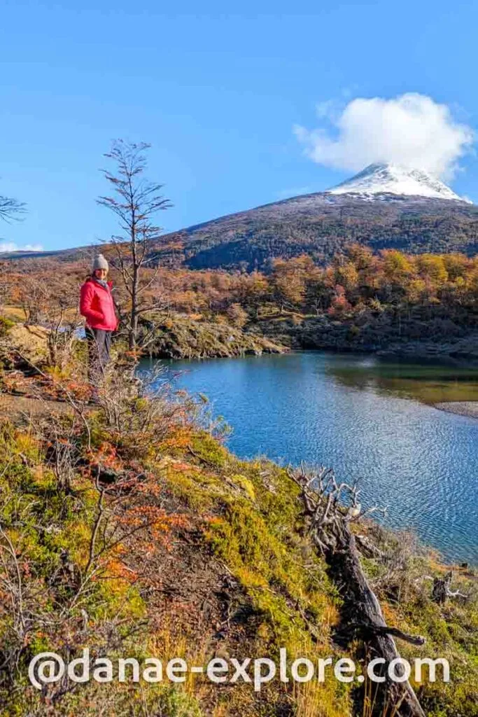 Young woman in a red coat and hat, amidst a landscape with autumn foliage, on the edge of a lake in Tierra del Fuego National Park. In the background is a snowy peak