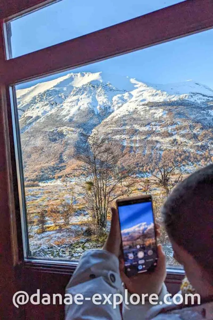Girl takes a photo with her cell phone through the window of the End of the World Train, there is a field and snowy mountains in the background