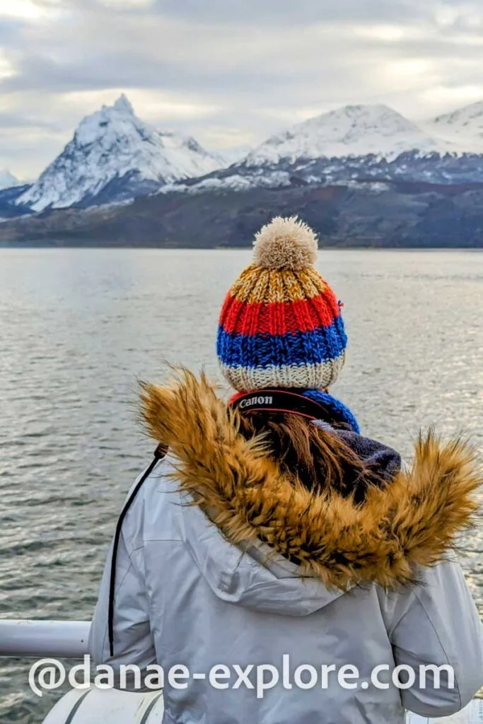 Girl in coat and hat, with her back to the camera, observes the landscape aboard a boat in the Beagle Channel. There are snow-capped mountains in the background