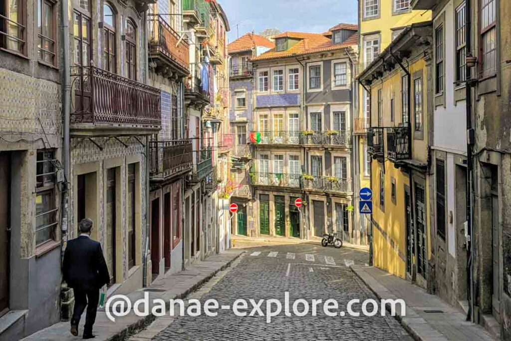 Homem andando na calçada em rua do Porto, Portugal. A rua é de paralelepípedos com prédios baixos com fachadas em azuleijo e sacadas em emtal