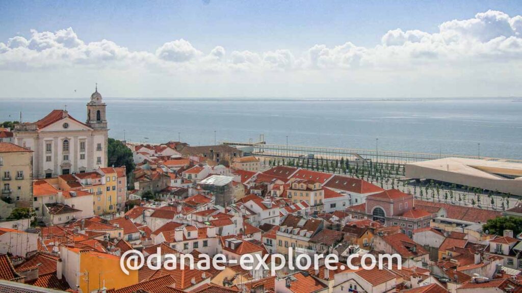 Cidade de Lisboa vista do Miradouro  de Santa Luzia, em um dia de sol e céu azul com poucas nuvens. Ao fundo se vê o Oceano Atlântico