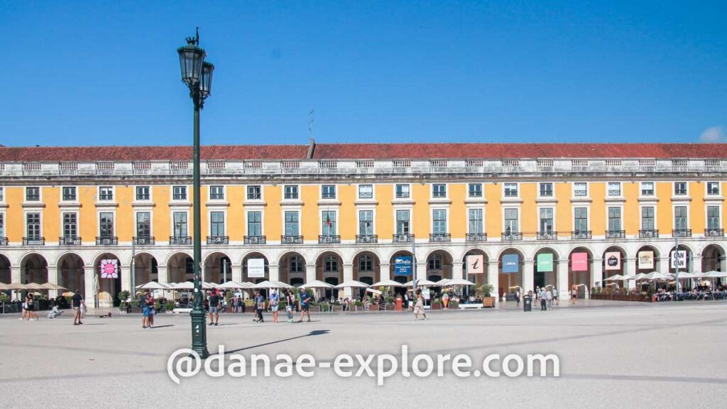 Praça do Comércio, em Lisboa, em um dia de verão com céu azul e sol. Na imagem, vê-se um prédio baixo de paredes amareladas e arcos no térreo, onde há guarda-sóis de restaurantes e pessoas caminhando.