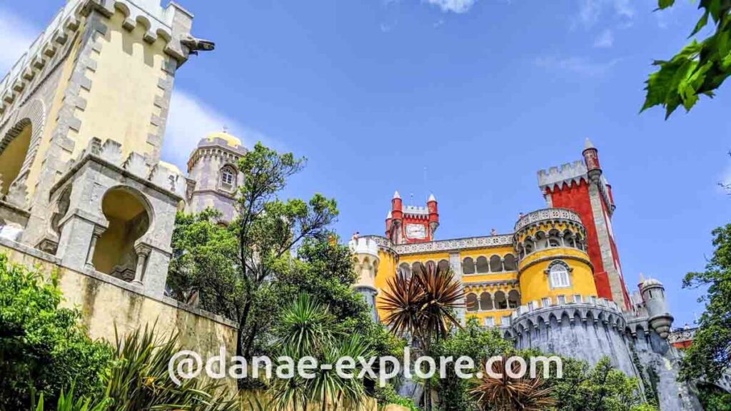 Palácio Nacional da Pena, em Sintra, Portugal. Se vêem os muros do castelo e suas paredes amarelas e vermelhas, em dia de sol e céu azul.