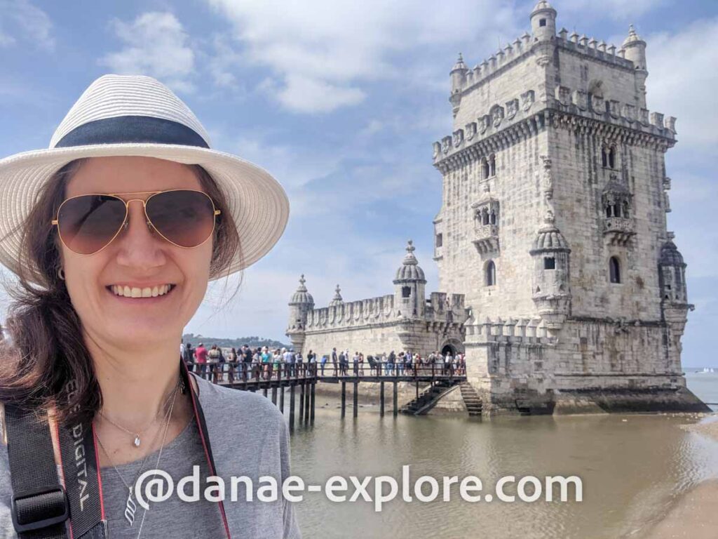 Eu, com óculos de sol e chapéu, em frente à Torre de Belém, em Lisboa, em dia de sol, céu azul com poucas nuvens. Lisboa foi a primeira parada em nosso roteiro de duas semanas em Portugal